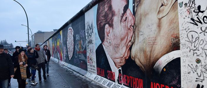 A painted stretch of the Berlin Wall at the East Side Gallery running away into the distance under a grey winter sky, the nearest segment carrying the mural of two men kissing on the mouth above red Cyrillic and hand-lettered German text, its surface covered in visitors' scribbled names; people in coats and scarves walk along the wet pavement beside it.