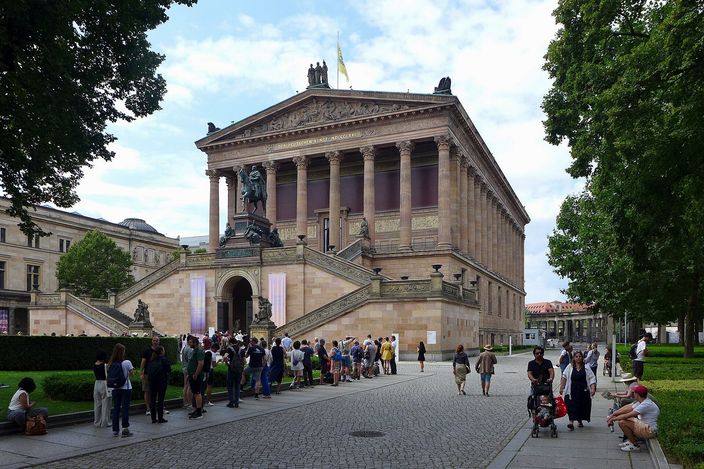 The Alte Nationalgalerie on Berlin's Museum Island in summer sunshine, a temple front of Corinthian columns raised on a high plinth above a double flight of steps, a queue of visitors waiting on the stairs and people walking on the tree-lined path in front.