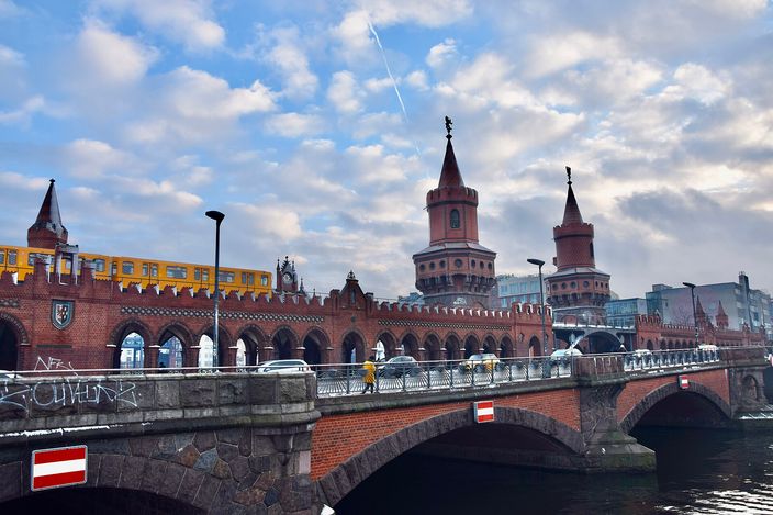 The Oberbaum Bridge in Berlin seen from the river: a double-decked red brick bridge with pointed twin towers at its centre, a yellow U-Bahn train crossing the arcaded upper deck, cars and pedestrians on the road below, under a sky of broken cloud.