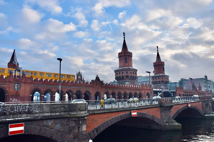 The Oberbaum Bridge in Berlin seen from the river: a double-decked red brick bridge with pointed twin towers at its center, a yellow U-Bahn train crossing the arcaded upper deck, cars and pedestrians on the road below, under a sky of broken cloud.