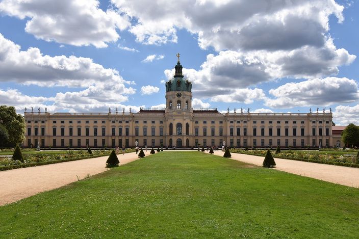 The garden front of Charlottenburg Palace in Berlin under a blue sky with white cloud: a long two-storey baroque range with a green copper dome and gilded weathervane over its centre, a mown lawn and gravel paths in front, and clipped conical topiary in the parterre.