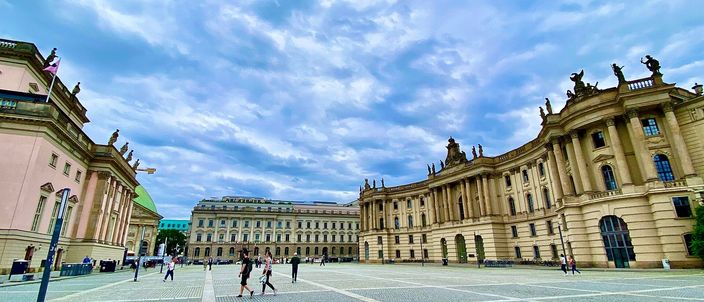 Bebelplatz in Berlin under a tall cloudy sky: the colonnaded Staatsoper on the left with statues along its roofline, the green dome of St Hedwig's Cathedral behind it, and the curved baroque facade of the old royal library on the right, with a few people crossing the open paved square between them.