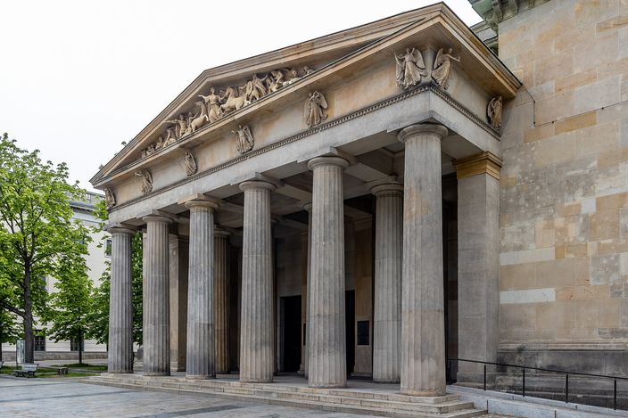 The Neue Wache in Berlin: a small classical guardhouse with six fluted Doric columns supporting a pediment carved with a battle relief, its single doorway standing open and dark, seen in soft daylight with trees to the left and the pale stone flank of the neighbouring arsenal to the right.