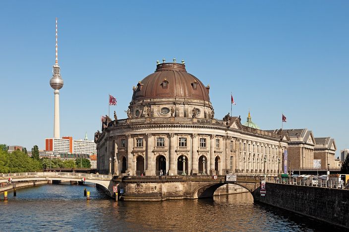The Bode Museum seen across the Spree on a clear summer day: a domed neo-baroque building standing on the rounded northern point of Museum Island like the bow of a ship, its brown dome and colonnaded flank above the water, the Berlin television tower rising behind it on the left.