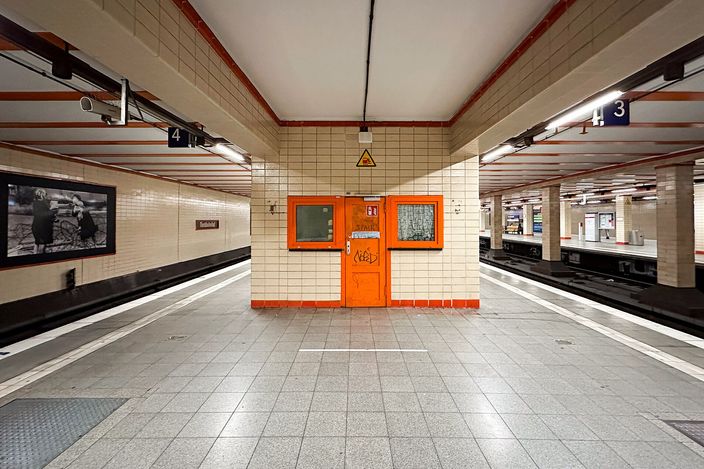 An empty tiled underground platform hall at Berlin Nordbahnhof station: cream tiles, an orange painted door in the central wall, platform numbers 3 and 4 on hanging signs, the old station nameplate on the left wall, and beside it a large framed black-and-white photograph from the years of the divided city showing figures at a barbed-wire barrier.