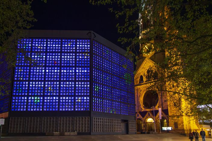 The Kaiser Wilhelm Memorial Church in Berlin at night: on the left Egon Eiermann's modern church, a rectangular block whose walls are made entirely of small glass squares glowing deep blue from within, and on the right the floodlit sandstone ruin of the old church tower with its lit clock face and gothic arches, seen from the square with a few people walking past.