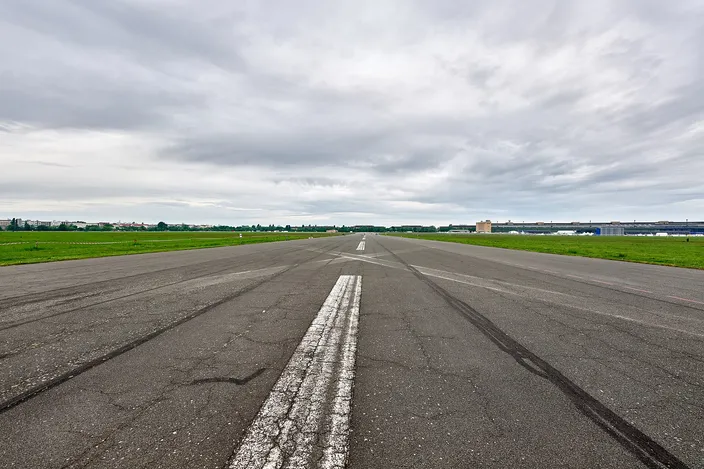 A disused airport runway at Tempelhofer Feld in Berlin photographed from ground level along its centreline: cracked gray asphalt with a broken white line running dead straight to a distant horizon, flat mown grass on both sides, the long low curve of the old terminal building along the right-hand edge, under a heavy overcast sky.