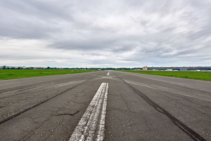 A disused airport runway at Tempelhofer Feld in Berlin photographed from ground level along its centreline: cracked grey asphalt with a broken white line running dead straight to a distant horizon, flat mown grass on both sides, the long low curve of the old terminal building along the right-hand edge, under a heavy overcast sky.
