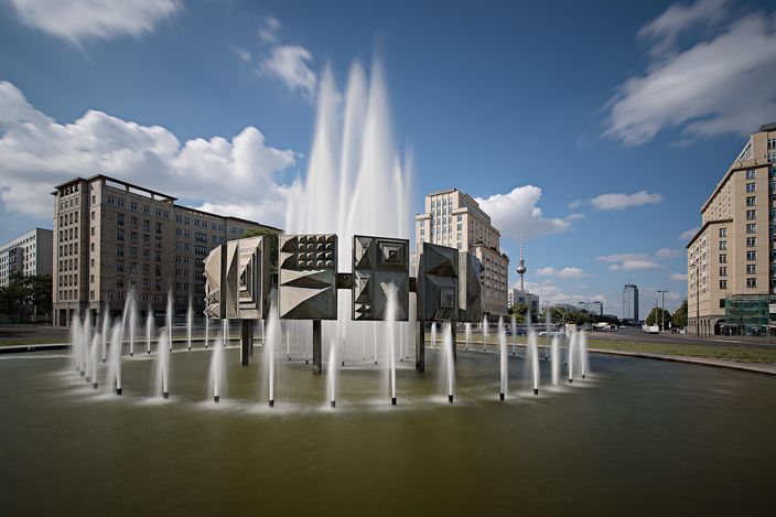 The fountain at Strausberger Platz in Berlin on a sunny day: a ring of water jets around a large abstract sculpture of geometric metal panels, with a tall central jet, framed left and right by the cream-tiled apartment blocks of Karl-Marx-Allee and with the television tower visible in the gap behind.