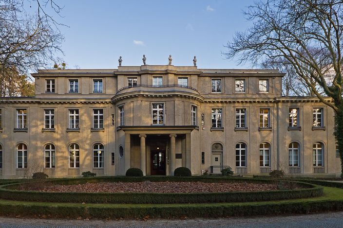 The House of the Wannsee Conference: a symmetrical two-storey villa in pale render with tall arched ground-floor windows, a rounded central bay above a small columned porch, and stone figures along the roofline, seen head-on across a gravel forecourt and a low clipped hedge in bare winter trees.