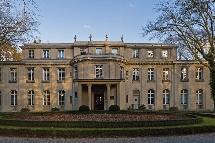 The House of the Wannsee Conference: a symmetrical two-story villa in pale render with tall arched ground-floor windows, a rounded central bay above a small columned porch, and stone figures along the roofline, seen head-on across a gravel forecourt and a low clipped hedge in bare winter trees.
