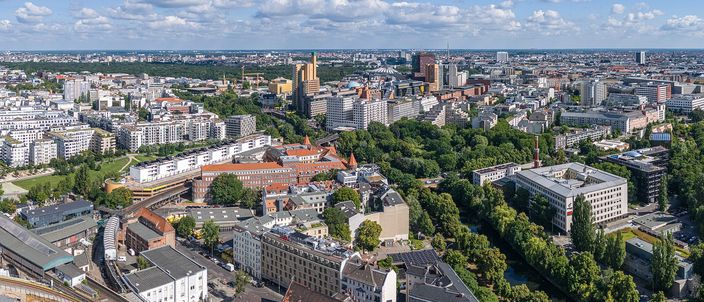 A high aerial view over Berlin on a summer afternoon: in the foreground an elevated railway viaduct with a yellow U-Bahn train on it, red-brick museum buildings and a wide green park beyond, a canal running away to the right, and in the middle distance the cluster of Potsdamer Platz towers under a big sky of scattered cumulus, with the flat city spreading unbroken to the horizon.