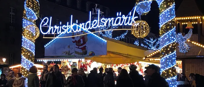 The lit gate of Strasbourg's oldest Christmas market at night, the word Christkindelsmärik in white lights across it, crowds and lit stalls beneath