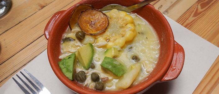 A wide bowl of ajiaco: a thick pale chicken-and-potato soup with a piece of corn on the cob, potato and slices of avocado.