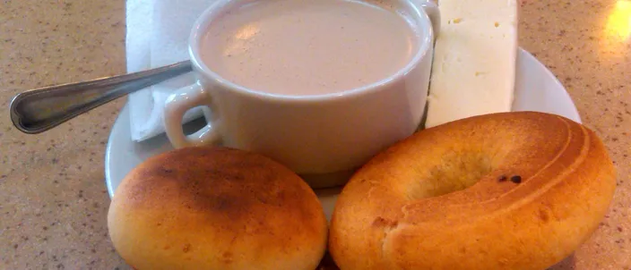 Two round golden almojábanas on a saucer beside a cup of hot chocolate with a slab of white cheese