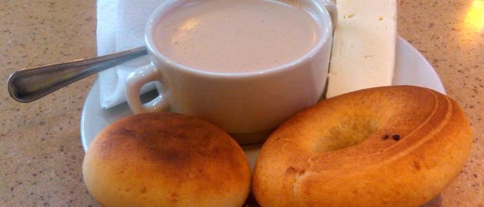 Two round golden almojábanas on a saucer beside a cup of hot chocolate with a slab of white cheese