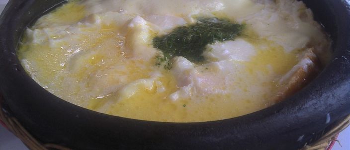 A black clay bowl of changua: a milky soup with a poached egg and softened bread, topped with chopped fresh green herbs, sitting in a woven basket.