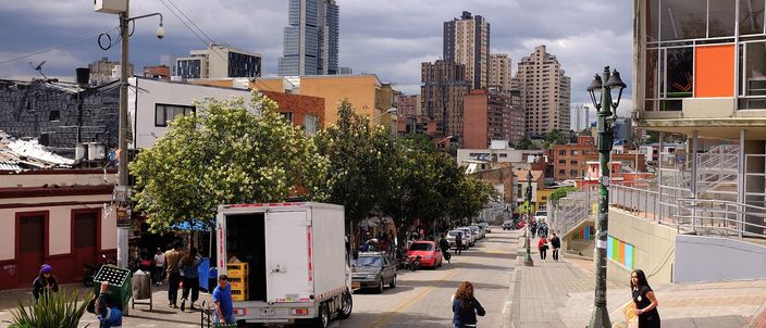 A Bogotá street sloping down towards the mountains under a grey sky, with a delivery truck, workers in blue overalls, pedestrians and a painted yellow zebra crossing in the foreground, low colonial-era buildings mixed with mid-rise blocks, and a glass supertall tower rising in the mid-distance