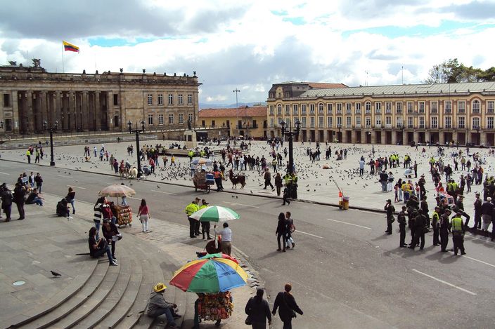 A very large paved square seen from a raised terrace, crowded with people and pigeons, a colonnaded neoclassical building flying the Colombian flag on the left and a long two-storey arcaded palace across the back, with vendors under bright umbrellas on the steps in the foreground
