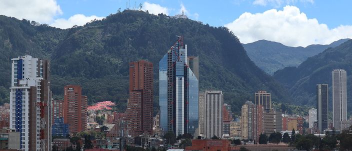 The green Monserrate ridge rising behind central Bogotá under cloud, with a small white building on the summit, and below it a dense band of red-brick mid-rise blocks and glass office towers