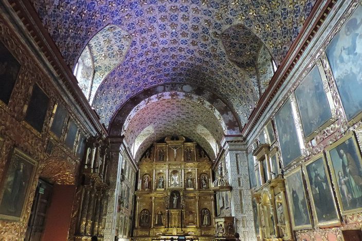 The single nave of the Iglesia Museo Santa Clara in Bogotá, its barrel vault covered end to end in gilded wooden flowers on deep blue, a tall gilded altarpiece of carved saints at the far end and framed colonial paintings covering both side walls from floor to vault