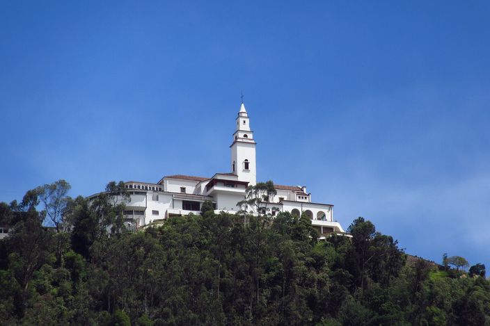 The white church of Monserrate with its slim bell tower standing on the wooded crest of a ridge against a deep blue sky, seen from below with dense green forest falling away beneath it