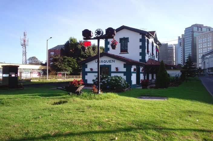 The small white-and-green wooden station house at Usaquén in Bogotá, its name painted across the gable, with flowerbeds and a period railway signal post with lamps in front of it, a lawn in the foreground and modern office towers behind