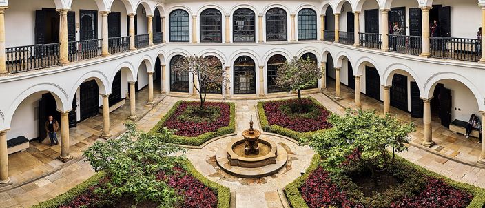 The two-storey arcaded courtyard of a white colonial house in Bogotá, with a stone fountain at the centre of four parterres of dark red and green planting, arched galleries on all four sides and a tiled roof against a bright sky