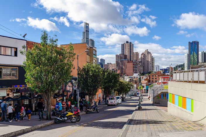 A Bogotá street in Santa Fe near La Candelaria: low shops and trees on the left with people gathered, a downhill street of mid-rise apartment blocks and a slim glass skyscraper under a blue sky with cumulus cloud.