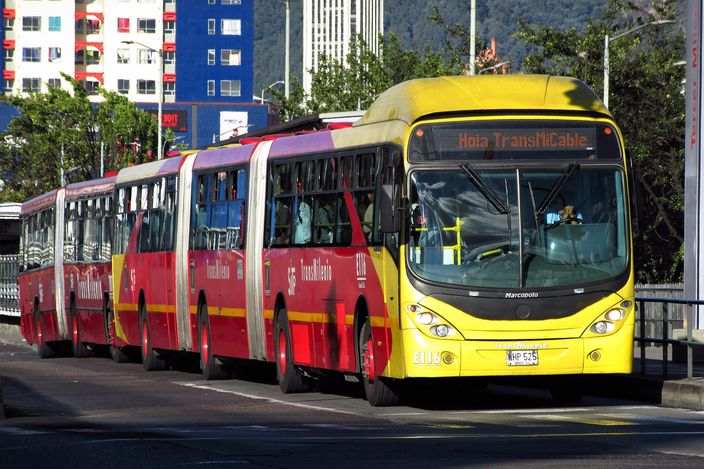 A red-and-yellow bi-articulated TransMilenio bus, destination sign reading 'Hola TransMiCable', on Avenida Caracas with apartment blocks and the green cerros orientales behind