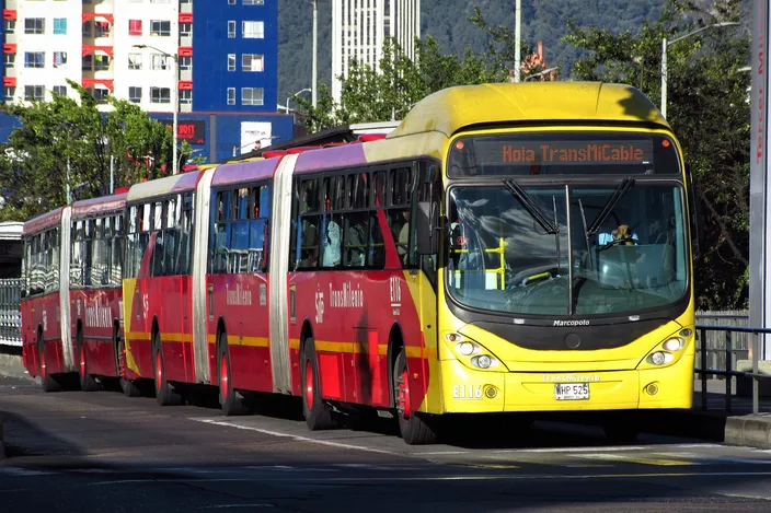 A red-and-yellow bi-articulated TransMilenio bus, destination sign reading 'Hola TransMiCable', on Avenida Caracas with apartment blocks and the green cerros orientales behind