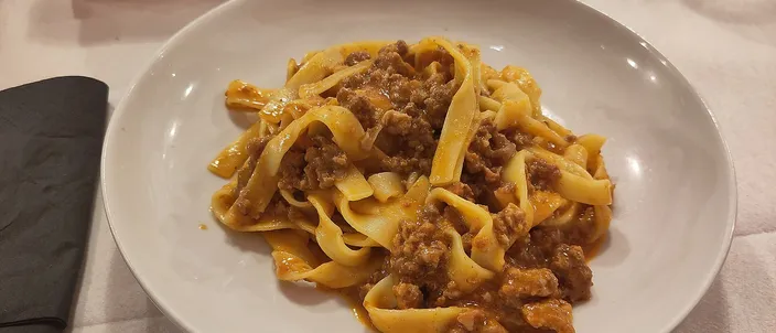 A plate of tagliatelle al ragù on a white tablecloth, the hand-cut ribbons evenly coated in a brown, meat-forward sauce with no pool of red on top.