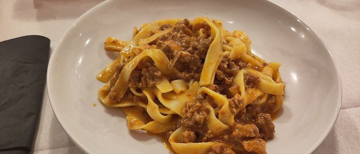 A plate of tagliatelle al ragù on a white tablecloth, the hand-cut ribbons evenly coated in a brown, meat-forward sauce with no pool of red on top.