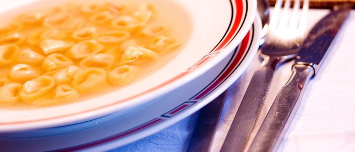 A shallow bowl of tortellini floating in clear golden broth, set at a laid table with a fork and knife