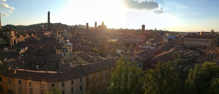 Bologna's terracotta rooftops at sunset, seen from above, with the city's medieval brick towers breaking the skyline and low hills behind.