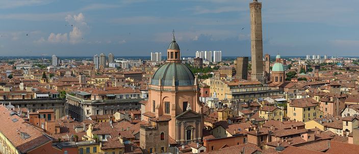 Bologna's terracotta rooftops in summer sunlight, with a domed church in the foreground and the tall Asinelli tower rising behind it, the hills and modern blocks of the outskirts on the horizon.