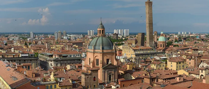 Bologna's terracotta rooftops in summer sunlight, with a domed church in the foreground and the tall Asinelli tower rising behind it, the hills and modern blocks of the outskirts on the horizon.