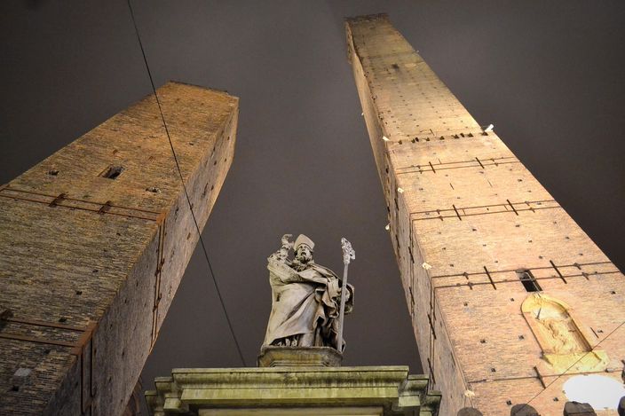 Bologna's two leaning medieval towers, the taller Asinelli and the shorter Garisenda, seen from below with a statue of San Petronio in the foreground.