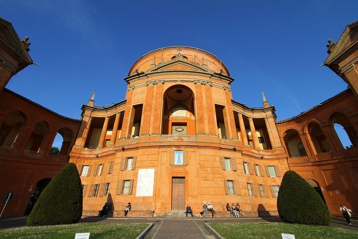 The Baroque Sanctuary of the Madonna di San Luca on the Colle della Guardia above Bologna: a terracotta basilica with a dome and an arched portico entrance, under a blue sky.