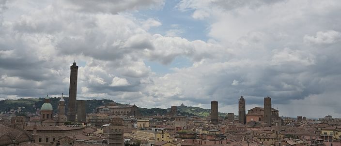 Bologna's terracotta rooftops under a broken grey sky, with the city's two leaning medieval towers and a church dome rising above the roofline.