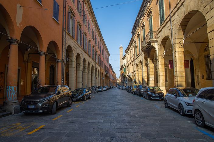 Strada Maggiore in Bologna: ochre and terracotta palaces with arcaded porticoes line both sides of a cobbled street, and one of the city's medieval brick towers closes the view at the far end.