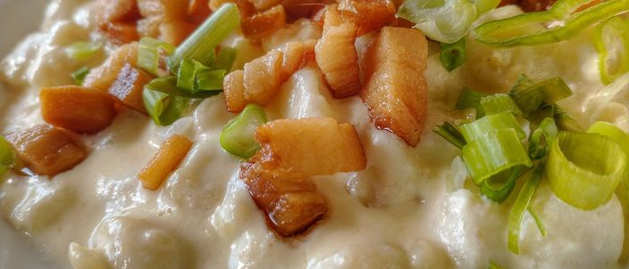 A close plate of small soft potato dumplings coated in white sheep's-milk bryndza, topped with cubes of crisp fried bacon and chopped spring onion.