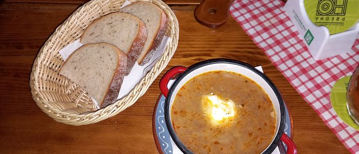 A rustic red enamel pot of dark, thick sauerkraut soup topped with a dollop of sour cream, set on a wooden table beside a basket of sliced bread.