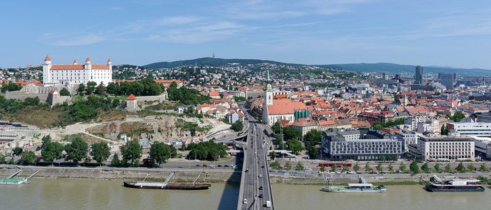 Panorama over the Danube with Bratislava Castle on its hill at left, St Martin's Cathedral spire and terracotta Old Town roofs at centre, and a road bridge crossing the river