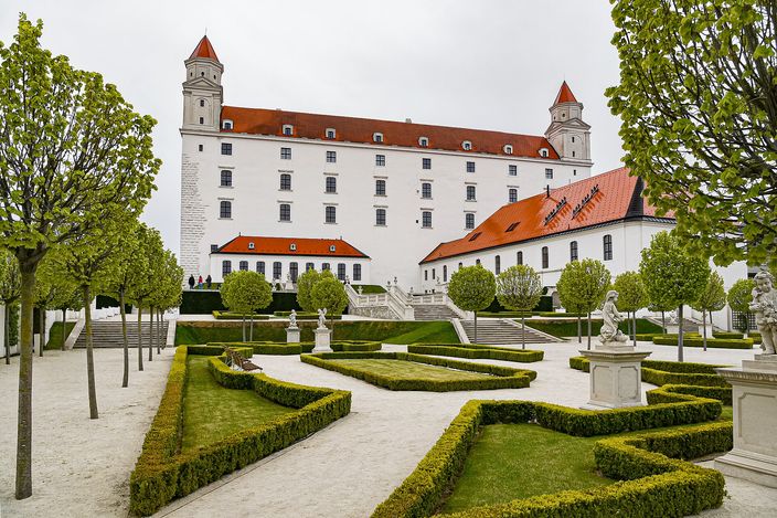 The white four-towered block of Bratislava Castle seen across its restored baroque garden, with clipped box hedges in geometric beds, gravel paths, stone statues and young trees under an overcast sky.