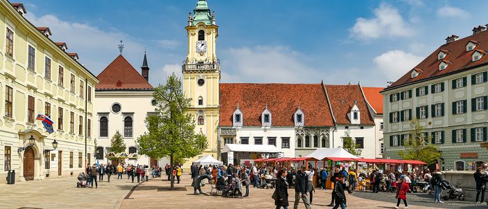 Bratislava's main square in spring sunshine: the Old Town Hall's tall pale clock tower with a green cupola at the centre, baroque houses closing the square on three sides, red-canopied market stalls under the arcades and people walking across the open paving.