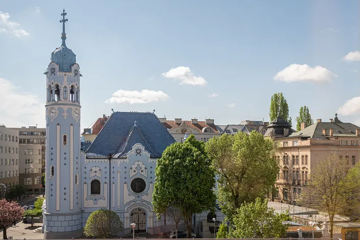A pale blue Art Nouveau church seen across an open space: a round blue bell tower with a clock on the left, a rounded gable and a blue glazed tiled roof at the center, green trees in front and ordinary apartment blocks closing the view behind.