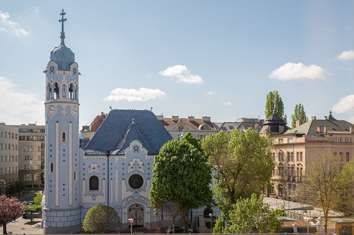 A pale blue Art Nouveau church seen across an open space: a round blue bell tower with a clock on the left, a rounded gable and a blue glazed tiled roof at the centre, green trees in front and ordinary apartment blocks closing the view behind.