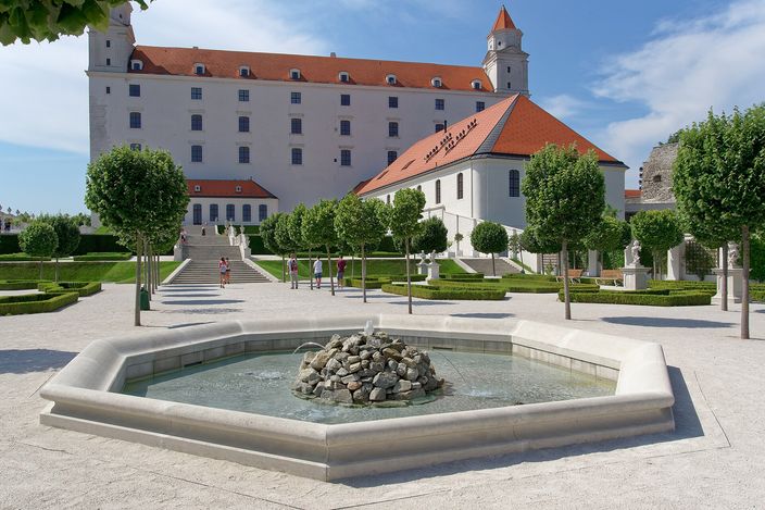 Bratislava Castle's white four-towered block on its terrace, seen across a formal garden of clipped trees and low box parterres with a stepped ramp climbing towards it and a low octagonal stone fountain basin in the foreground.