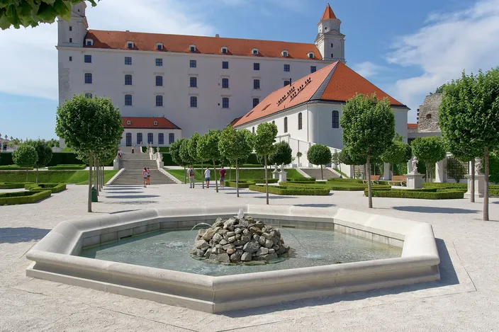 Bratislava Castle's white four-towered block on its terrace, seen across a formal garden of clipped trees and low box parterres with a stepped ramp climbing towards it and a low octagonal stone fountain basin in the foreground.